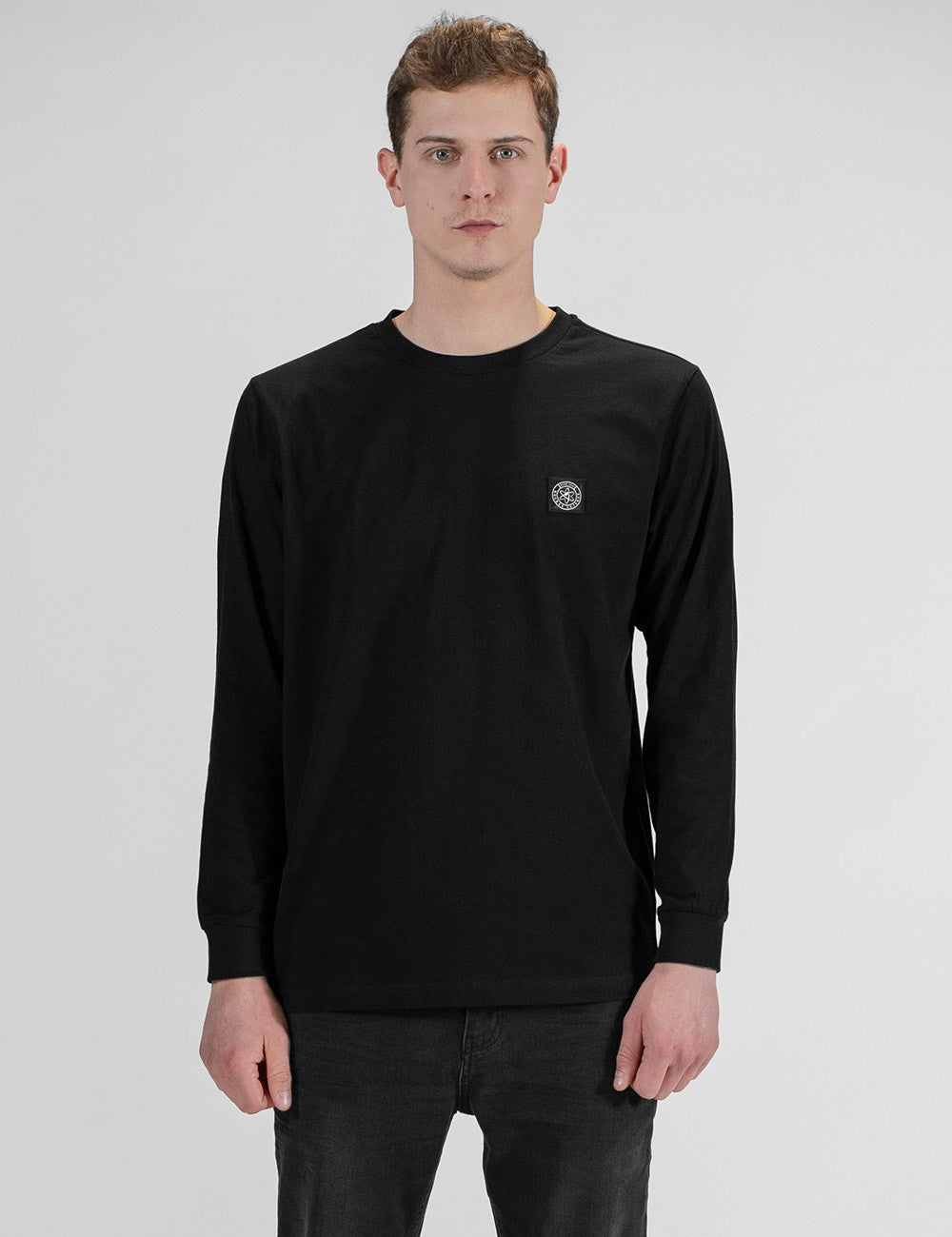 A front-facing shot of a model with short brown hair, wearing a long-sleeved black crewneck t-shirt with a small circular emblem on the left chest, paired with dark grey or black jeans
