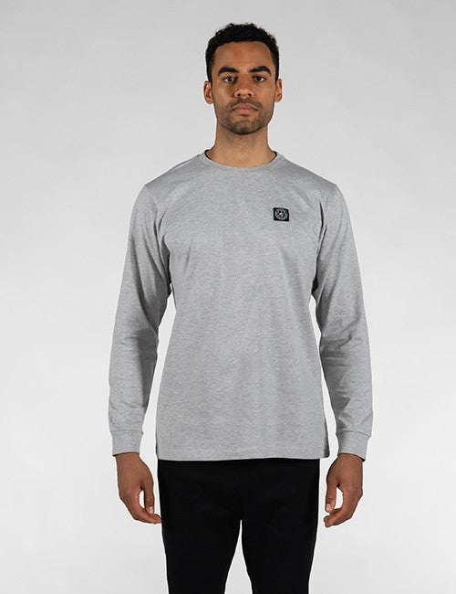 A man stands against a plain grey background, wearing a grey heather long-sleeve crewneck shirt with a small square black Rocket Science logo patch on the left chest and black pants.