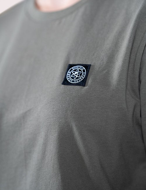 A close-up shot of the left chest area of a dark olive-green crewneck t-shirt. On the chest is a small, stitched square black patch containing a circular white emblem with an atomic symbol in the center.