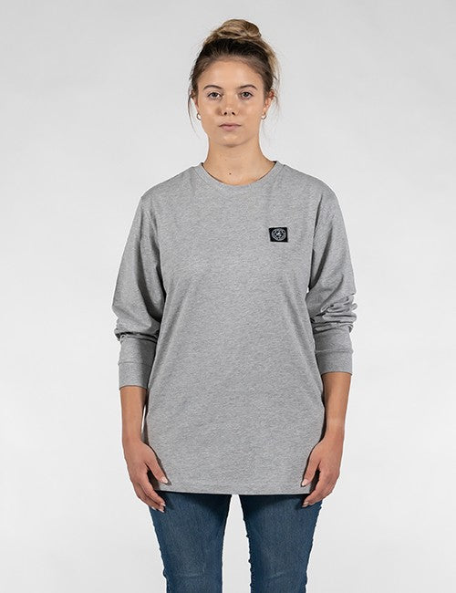 A woman modeling a heather gray long-sleeve crewneck sweatshirt with a small black square Rocket Science logo patch on the left chest, paired with blue jeans against a light gray studio background.