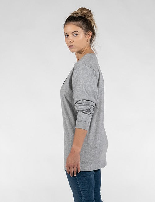 A profile shot of a woman with her hair in a messy bun, looking over her shoulder at the camera. She is wearing a grey long-sleeved t-shirt and blue jeans, posed against a solid light grey background.