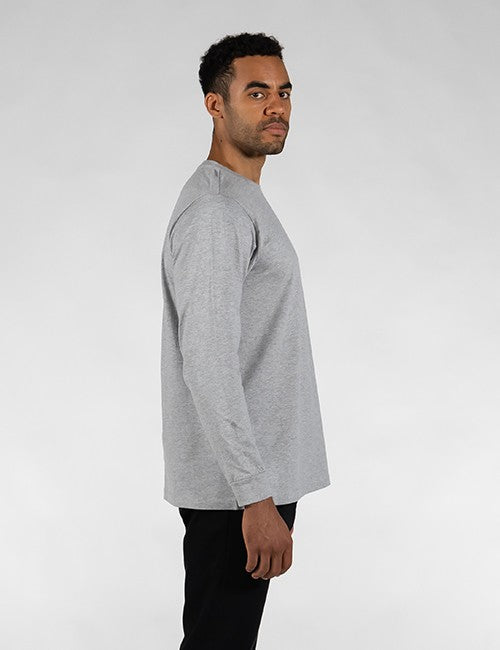 A side profile shot of a man with short black hair and a light beard, looking over his shoulder toward the camera. He is wearing a long-sleeved, heather grey crewneck t-shirt with a relaxed fit, paired with black pants, against a plain white background.