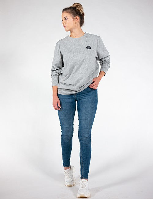 A full-length shot of a woman with her hair in a bun, wearing a grey long-sleeve crewneck sweatshirt, blue skinny jeans, and white sneakers.
