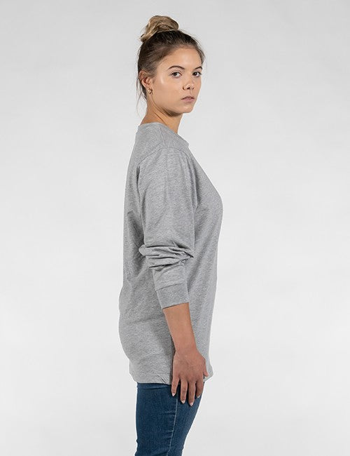 A woman with her hair in a high bun stands in a side-profile view against a plain, light grey background. She is wearing a loose-fitting, heather grey long-sleeve sweatshirt paired with blue jeans. Her right arm hangs naturally at her side, showcasing the ribbed cuff of the sleeve, while she turns her head slightly to look directly toward the camera with a neutral expression.
