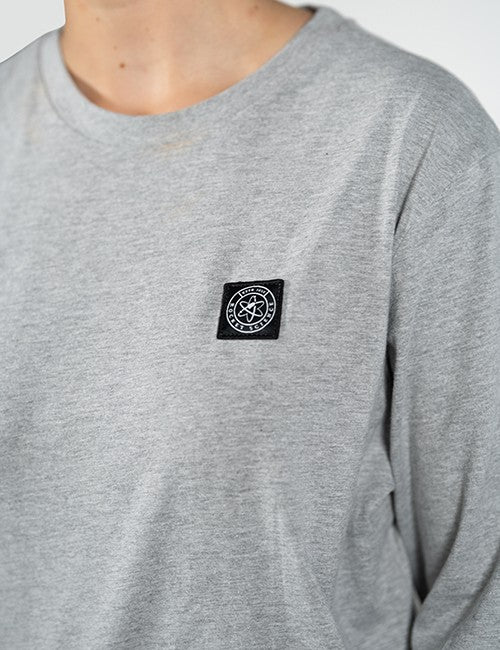 Close-up of a person wearing a heather gray long-sleeve t-shirt with a black square chest patch featuring a white circular Rocket Science logo.