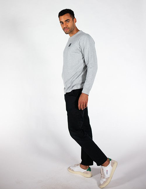 A man wearing a light gray long-sleeve crewneck sweatshirt, black pants, and white sneakers with green accents, standing in profile against a white studio background.