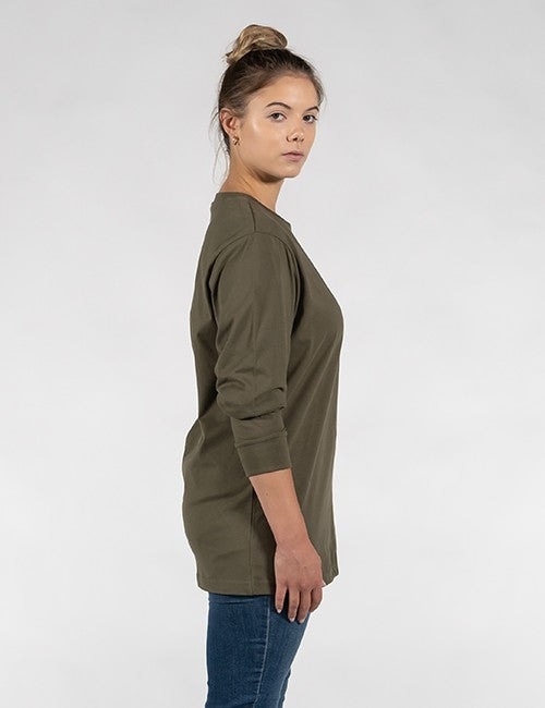 A young woman with light skin and brown hair tied in a high bun stands in a side-profile view against a plain, light-gray background. She is wearing an olive-green, long-sleeved crewneck t-shirt with ribbed cuffs and blue jeans, looking over her shoulder toward the camera.