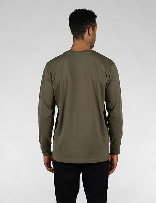 A man with short, dark curly hair is shown from the back against a plain light gray background. He is wearing a long-sleeved, solid olive-green shirt with ribbed cuffs and black pants.