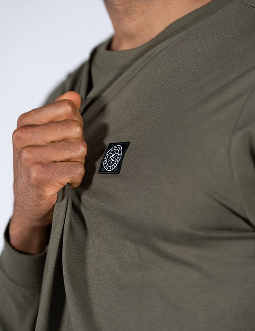 Close-up of a person wearing an olive green long-sleeve t-shirt, pulling the fabric to reveal a black rectangular patch with a white Rocket Science design on the chest.