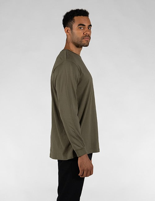 A man shown in profile wearing a long-sleeve crewneck shirt in olive green, paired with black pants, against a light gray studio background.