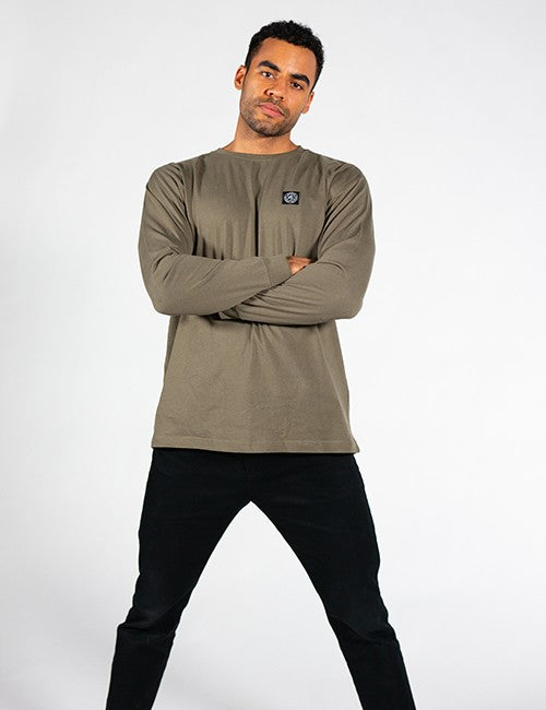 An athletic man with short, dark hair stands facing forward with his arms crossed over his chest. He is wearing an olive-green, long-sleeved t-shirt with a small square patch logo on the left chest, paired with black pants. The background is a solid, light gray.