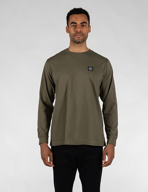 A man wearing an olive green long-sleeve crewneck shirt with a small circular Rocket Science logo patch on the left chest, paired with black pants, standing against a light gray studio background.