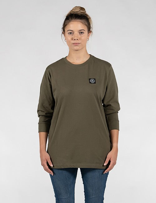 A woman modeling an olive green long-sleeve t-shirt with a small black rectangular logo patch on the left chest, paired with blue jeans, against a light gray studio background.