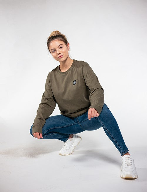 A young woman with blonde hair tied up in a messy bun is crouching forward against a plain white background. She is wearing a crewneck olive green long-sleeve sweatshirt featuring a small black patch logo on the left side of the chest, paired with slim-fit blue denim jeans and chunky white sneakers.