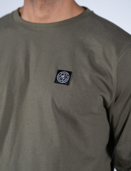 Close-up of a person wearing an olive green long-sleeve t-shirt with a black square Rocket Science logo patch on the left chest.
