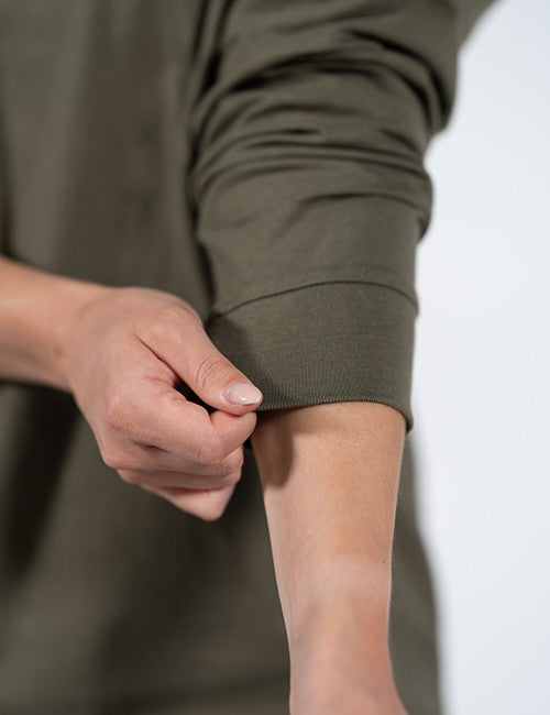 A close-up shot focuses on a person's forearm as they pull up the sleeve of an olive green long-sleeved shirt. The fabric is soft and slightly creased around the elbow, ending in a wide, ribbed cuff held by their fingers.