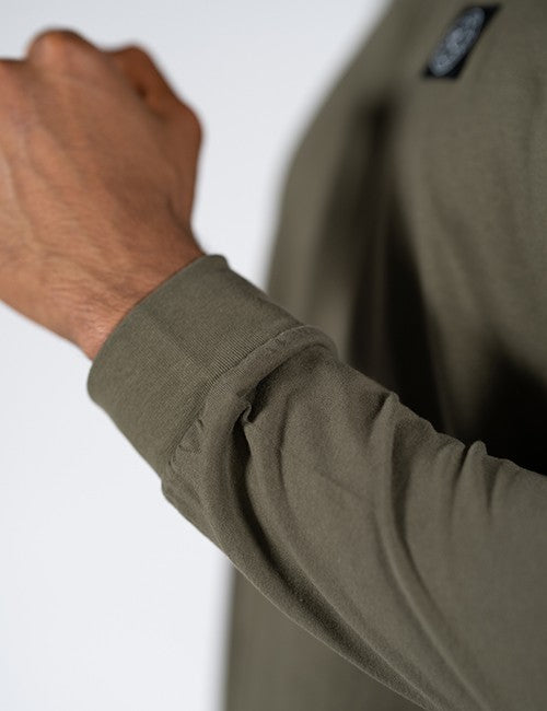 Close-up of a person wearing an olive green long-sleeve shirt, showing the sleeve cuff and a small black Rocket Science patch on the chest against a light gray background.