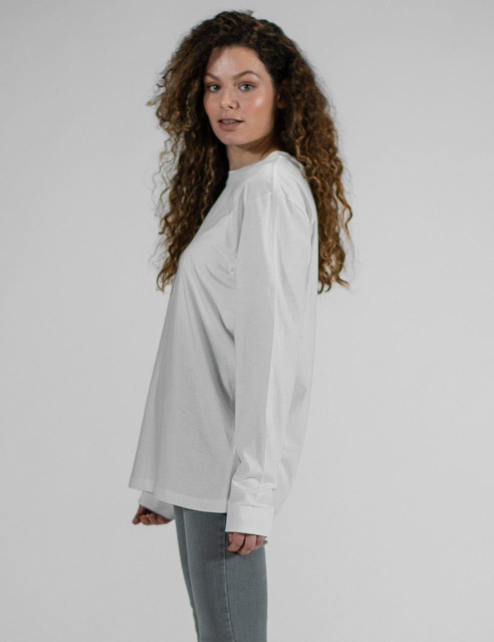 A female model with long, voluminous, curly brown hair stands in a side-profile view against a solid grey background. She is wearing a plain, loose-fitting white long-sleeved t-shirt with ribbed cuffs from Rocket Science and grey pants.