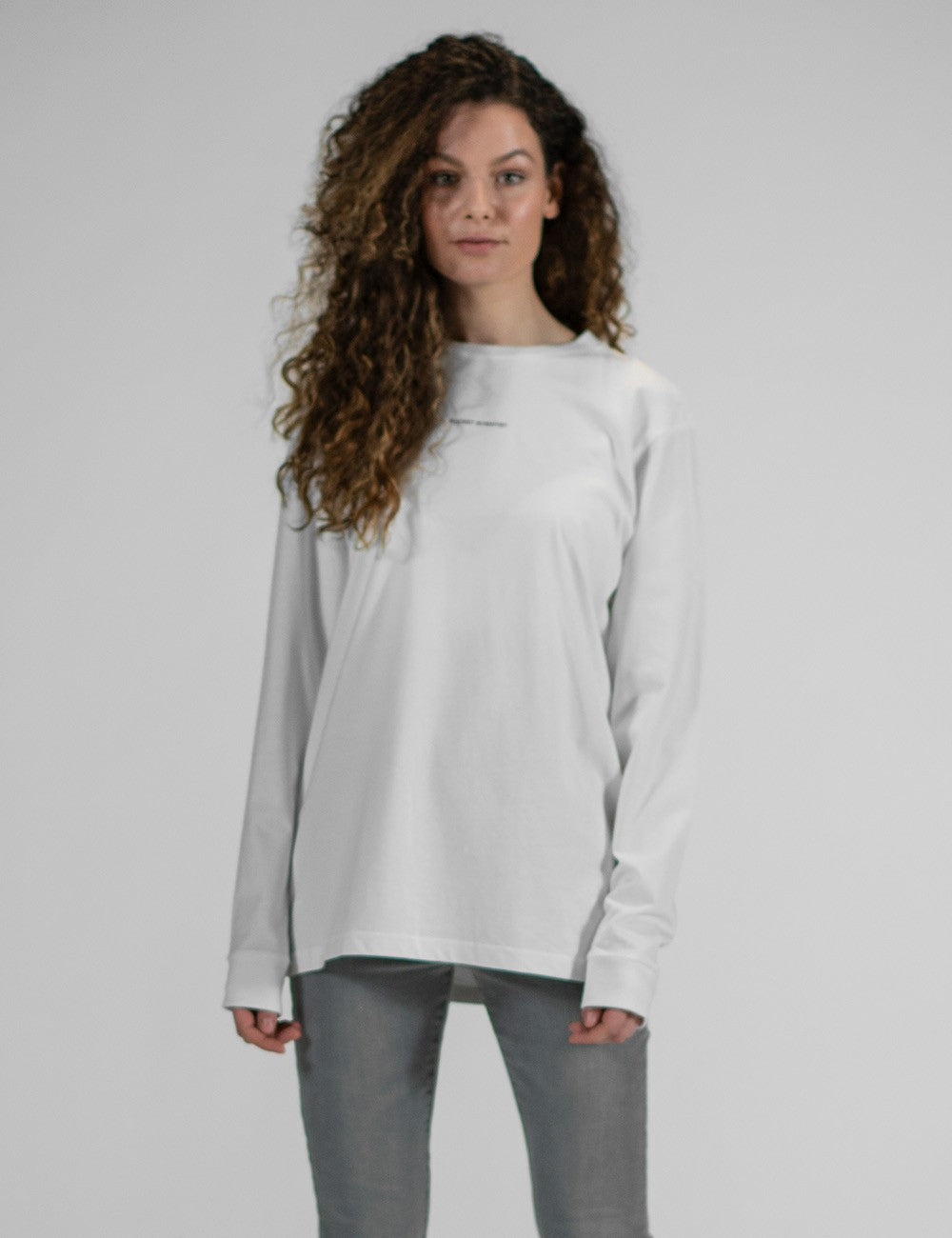 A woman with long, voluminous, curly brown hair stands in a front pose against a solid grey background. She is wearing a plain, loose-fitting white long-sleeved t-shirt with ribbed cuffs and grey pants.