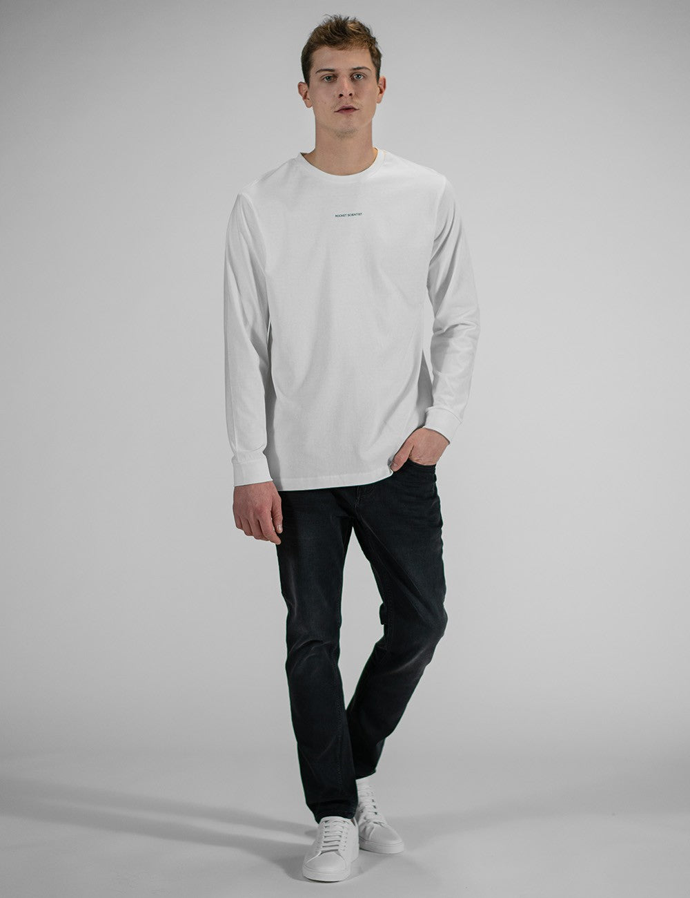 A young man wearing a white long-sleeve crewneck shirt with a small, minimalist "Rocket Science" logo printed in the center of the chest. He has paired the shirt with slim-fit dark navy or black jeans and clean white sneakers.