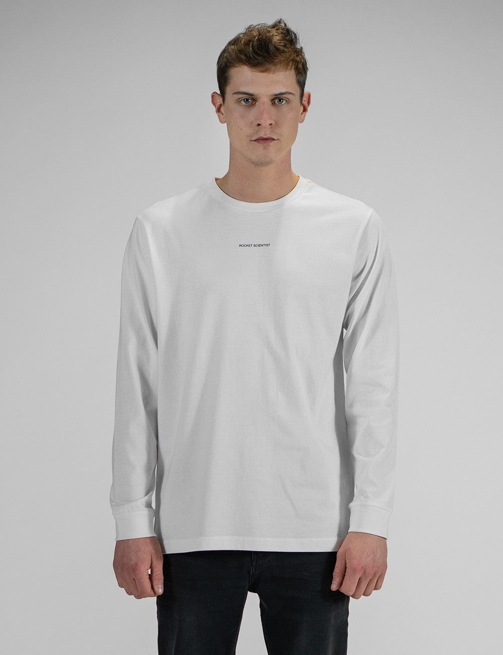 A young man wearing a white long-sleeve crewneck shirt with a small, minimalist "Rocket Science" logo printed in the center of the chest. He has paired the shirt with slim-fit black jeans.
