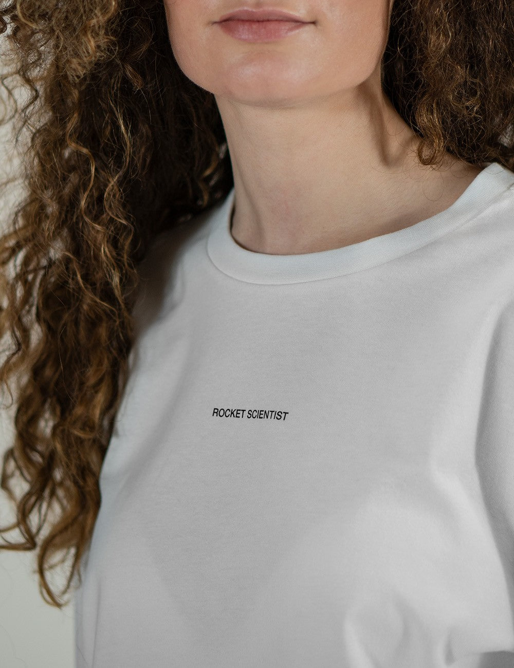 White crew neck longsleeve t-shirt featuring a small, minimalist text print that reads "ROCKET SCIENTIST" centered across the upper chest. The person wearing it has long, brown, curly hair draped over one shoulder.