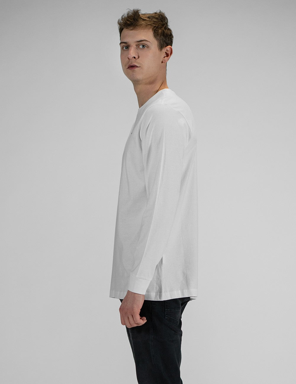 A young man wearing a white long-sleeve crewneck shirt with a small, minimalist "Rocket Science" logo printed in the center of the chest. He has paired the shirt with slim-fit black jeans.
