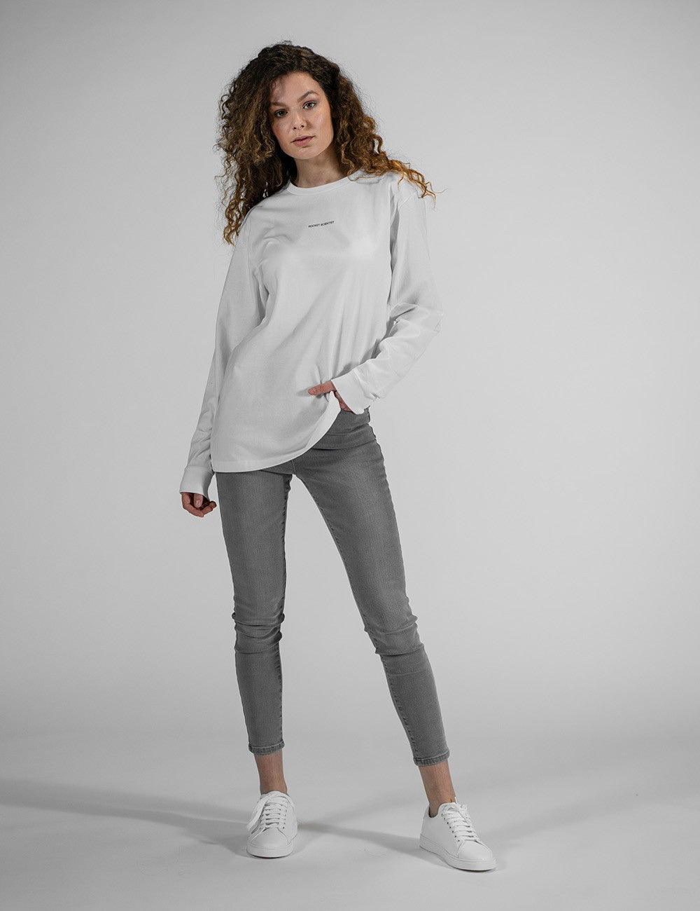 A full-length portrait of a female model with long, curly brown hair posing against a light gray background. She is wearing an oversized, long-sleeved white t-shirt, slim-fit gray jeans, and clean white sneakers. She stands with her feet split apart, leaning slightly to her left with her left hand resting gently near her hip pocket.
