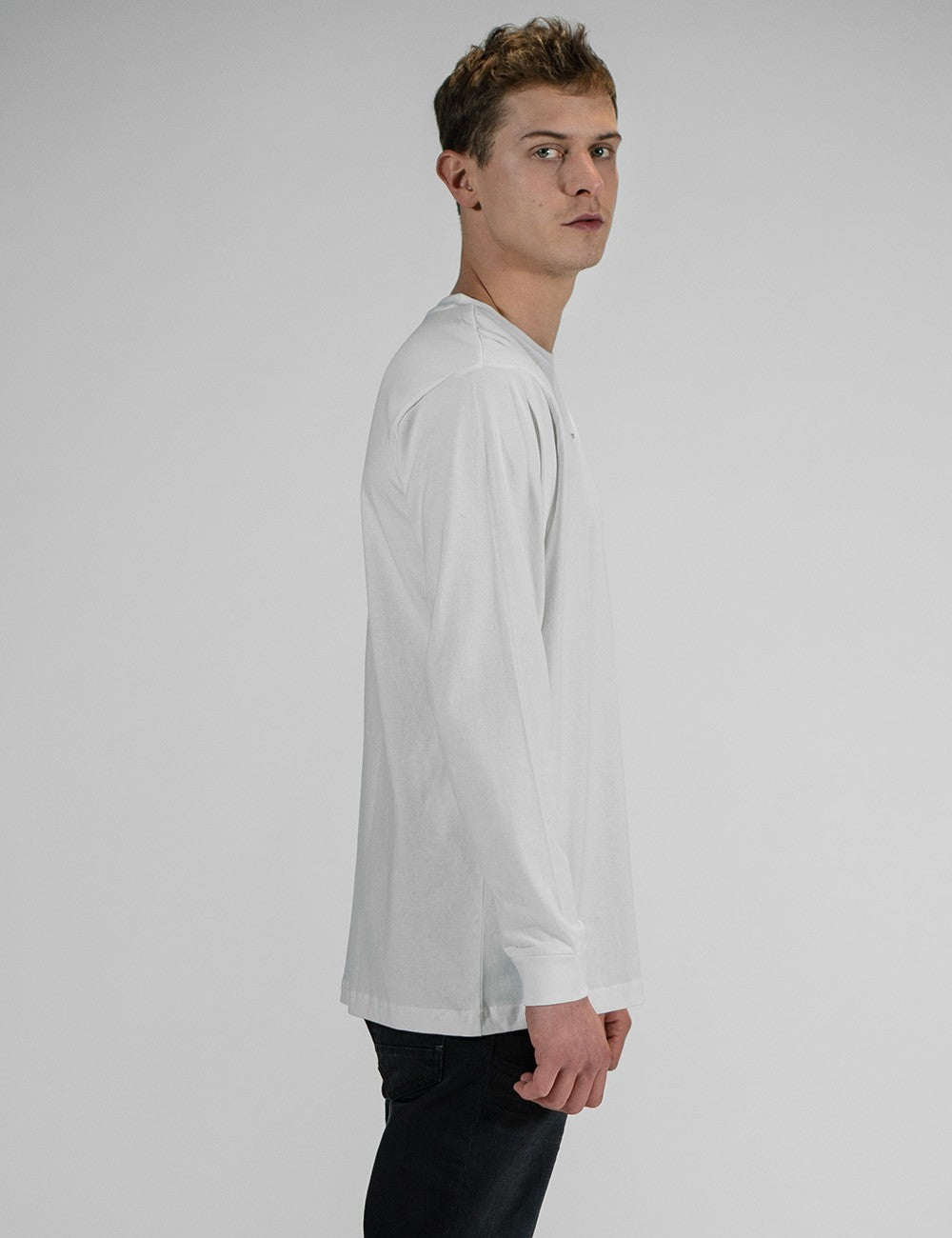 A young man wearing a white long-sleeve crewneck shirt with a small, minimalist "Rocket Science" logo printed in the center of the chest. He has paired the shirt with slim-fit  black jeans.