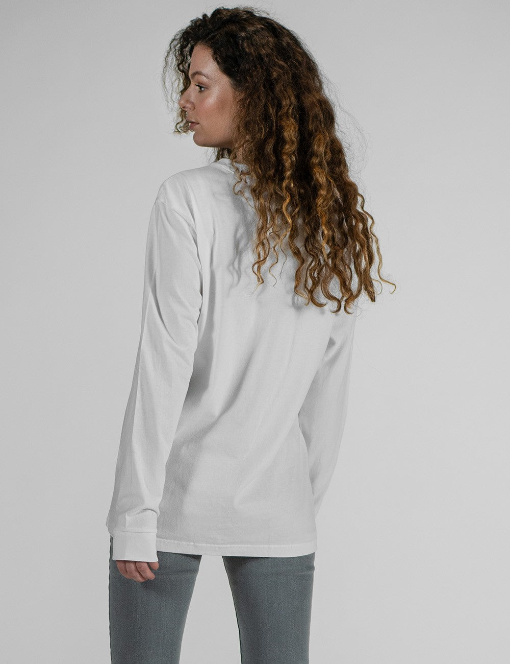 A female model with long, curly brown hair stands facing away and looking over her left shoulder against a plain grey background. She is wearing a plain white long-sleeve t-shirt and grey jeans.