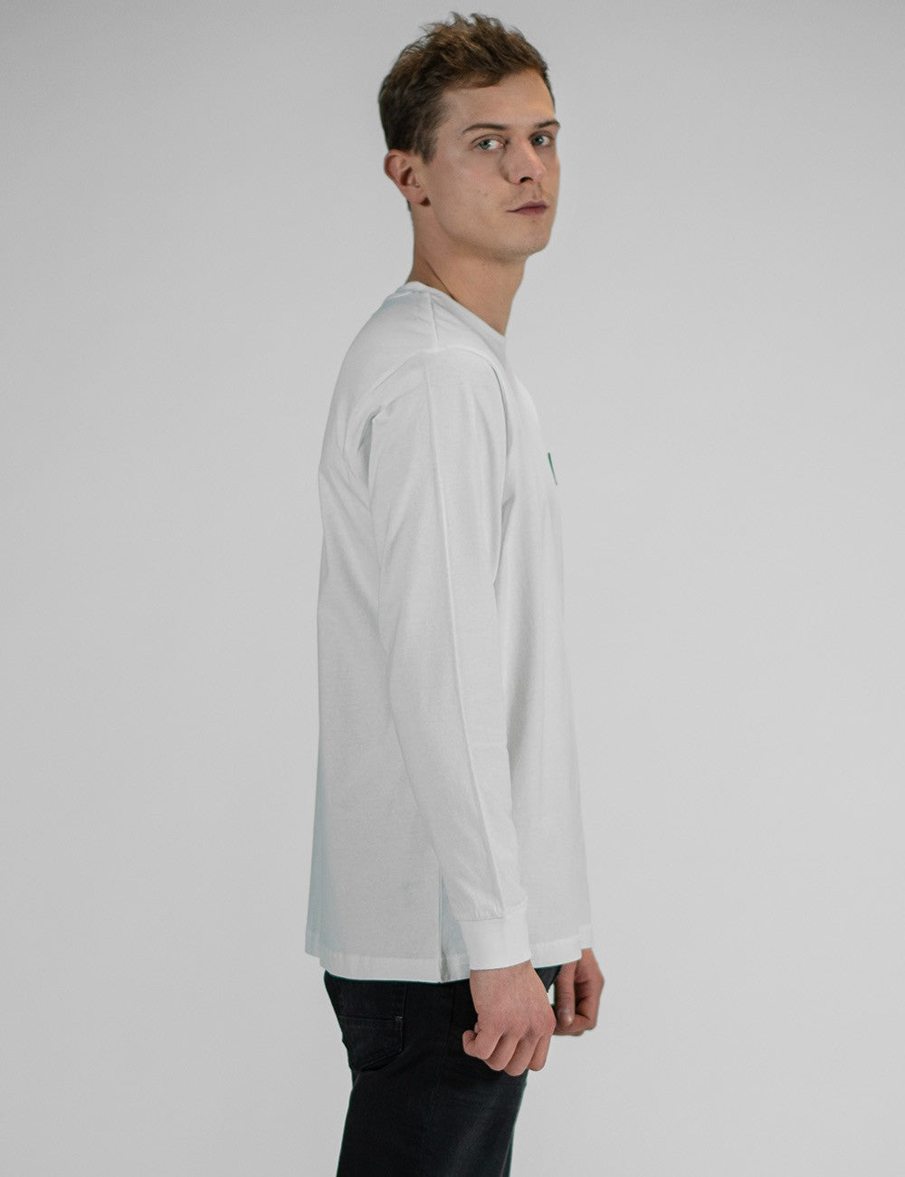 A person modeling the Rocket Science unisex white long-sleeve t-shirt with ribbed cuffs, featuring a small black logo patch on the left chest, styled casually with slim black jeans.