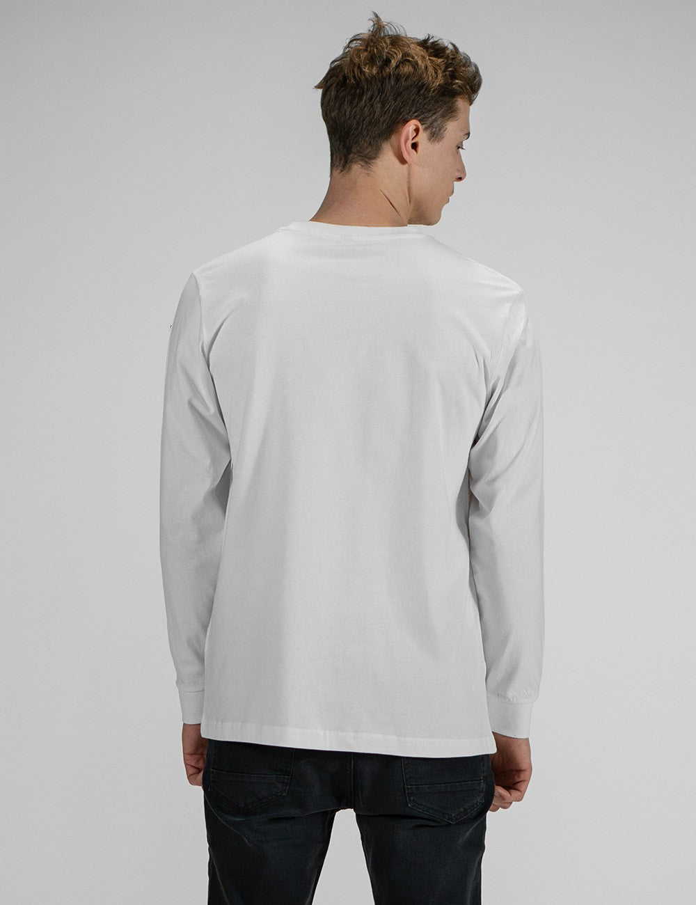 A person modeling the Rocket Science unisex white long-sleeve t-shirt with ribbed cuffs, featuring a small black logo patch on the left chest, styled casually with slim black jeans.