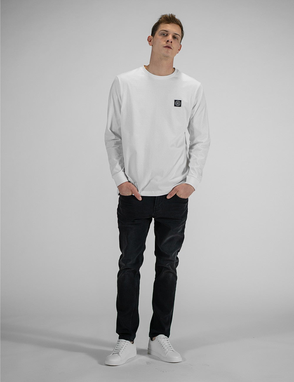 A person modeling the Rocket Science unisex white long-sleeve t-shirt with ribbed cuffs, featuring a small green and black logo patch on the left chest, styled casually with slim black jeans.