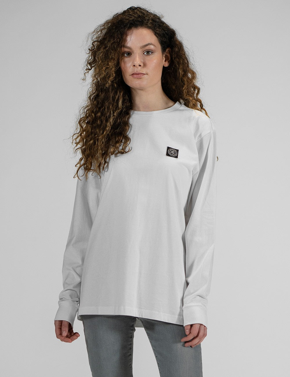 A person modeling the Rocket Science unisex white long-sleeve t-shirt with ribbed cuffs, featuring a small and black logo patch on the left chest, styled casually with slim grey jeans.