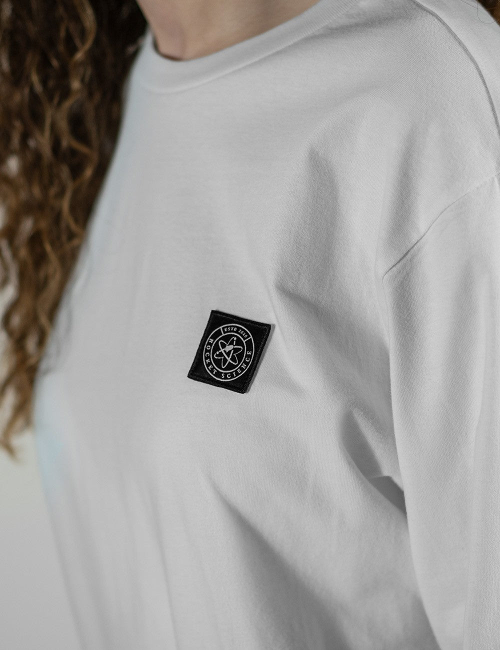 A close-up, side-profile view of a person wearing a plain white, long-sleeve crewneck shirt. The fabric has an oversized, dropped-shoulder fit. Attached to the left chest area is a small, square black patch featuring a circular embroidered emblem that contains an atom or rocket-like symbol with the text "Rocket Science" wrapped around the border. 