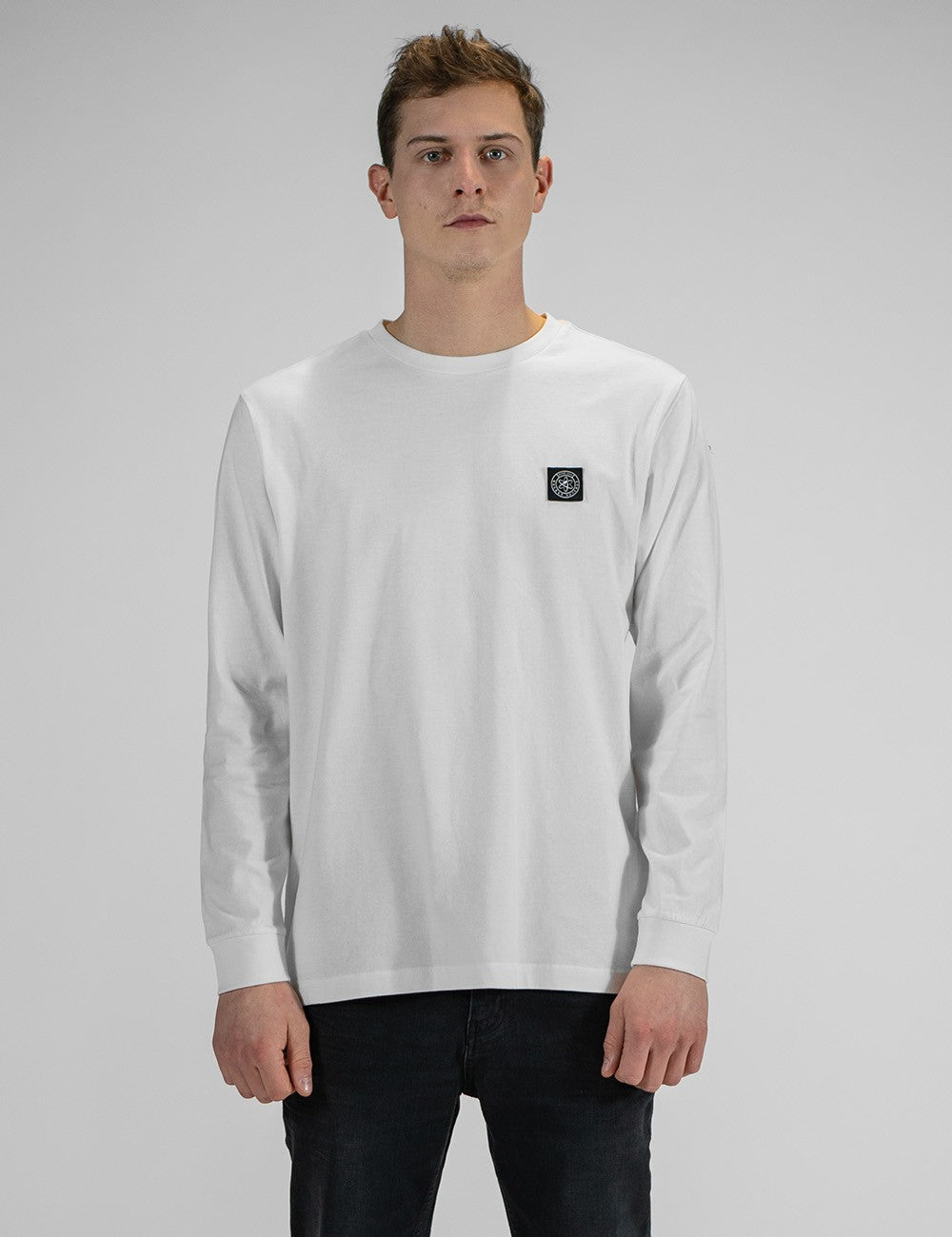 A person modeling the Rocket Science unisex white long-sleeve t-shirt with ribbed cuffs, featuring a small green and black logo patch on the left chest, styled casually with slim black jeans.