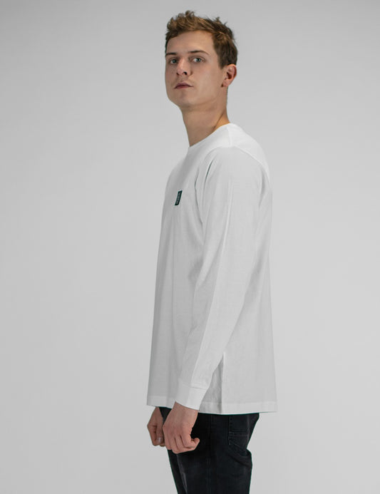 A person modeling the Rocket Science unisex white long-sleeve t-shirt with ribbed cuffs, featuring a small green and black logo patch on the left chest, styled casually with slim black jeans.