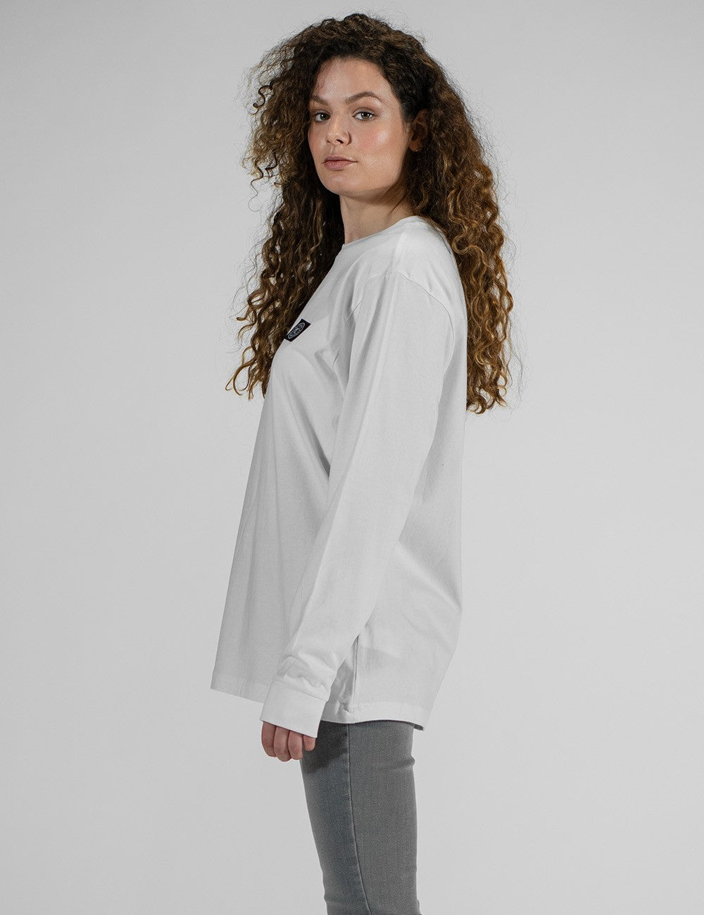 A person modeling the Rocket Science unisex white long-sleeve t-shirt with ribbed cuffs, featuring a small and black logo patch on the left chest, styled casually with slim grey jeans.