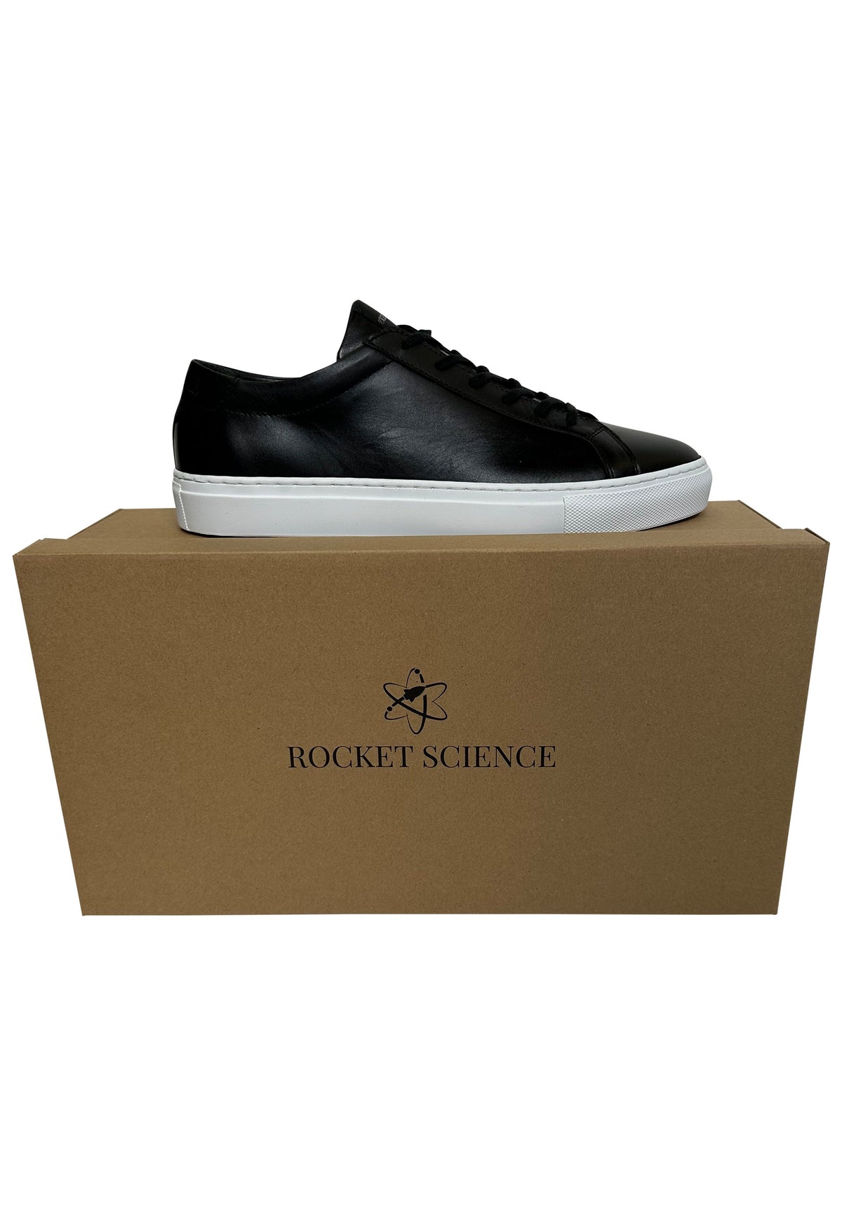 A black low-top sneaker with a leather upper and a thick white rubber sole sits on top of its plain cardboard shoe box, which features the "Rocket Science" logo and brand name printed in black ink on the front.