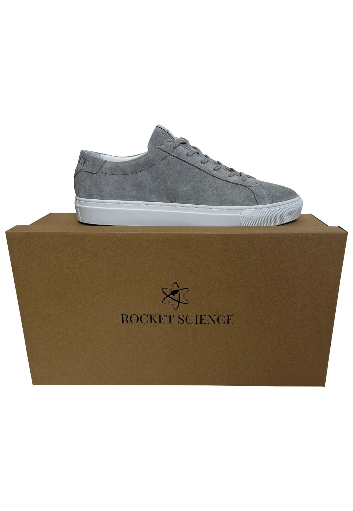 A grey suede low-top sneaker sits on top of its plain brown cardboard box. The shoe features matching grey laces and a solid white rubber sole. The front face of the box is stamped in black ink with the brand name "ROCKET SCIENCE" beneath an icon of an atom.