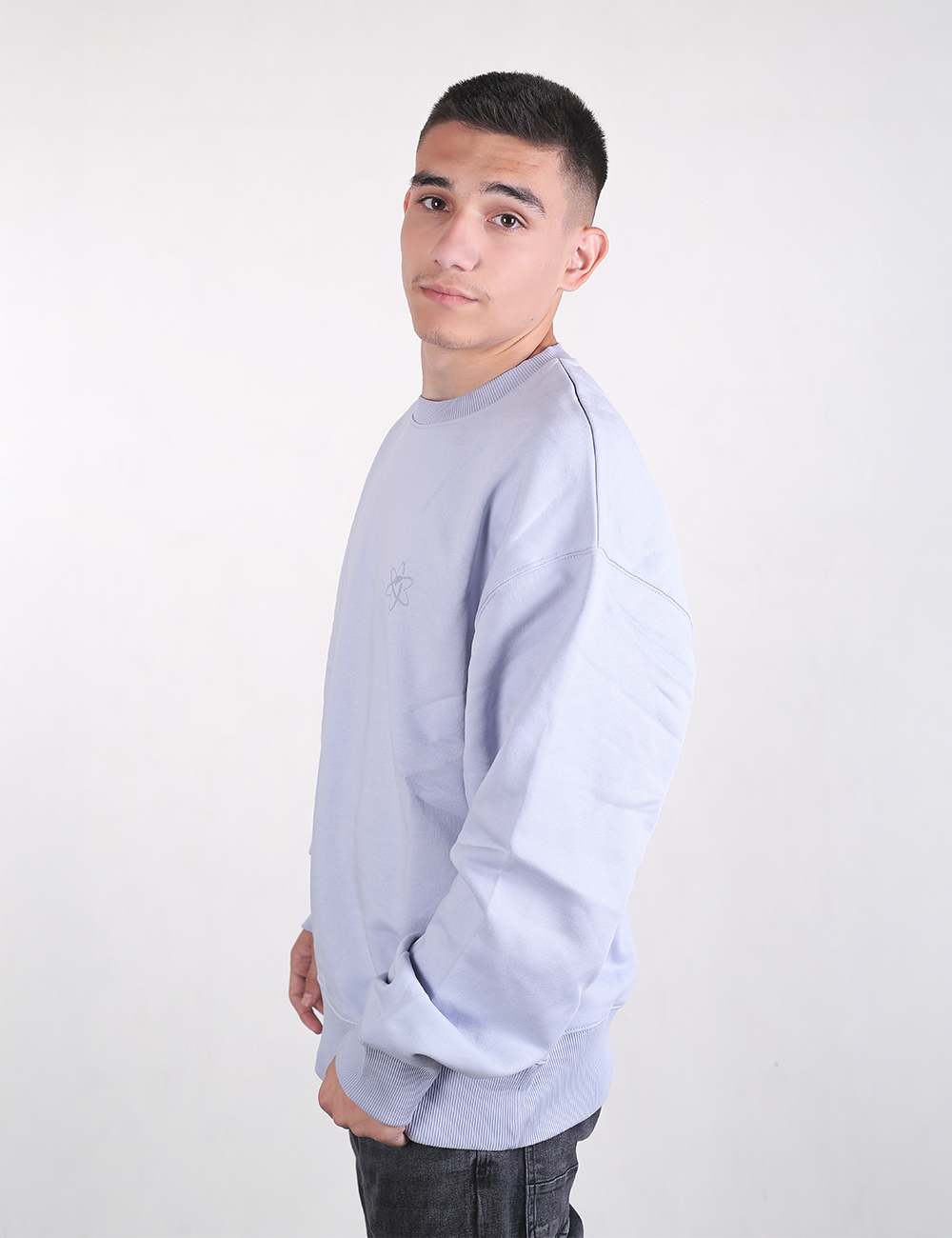 A profile view of a young man with short dark hair standing against a white backdrop. He is wearing a light lilac or pastel purple crewneck sweatshirt and dark jeans, looking directly at the camera with a neutral expression.