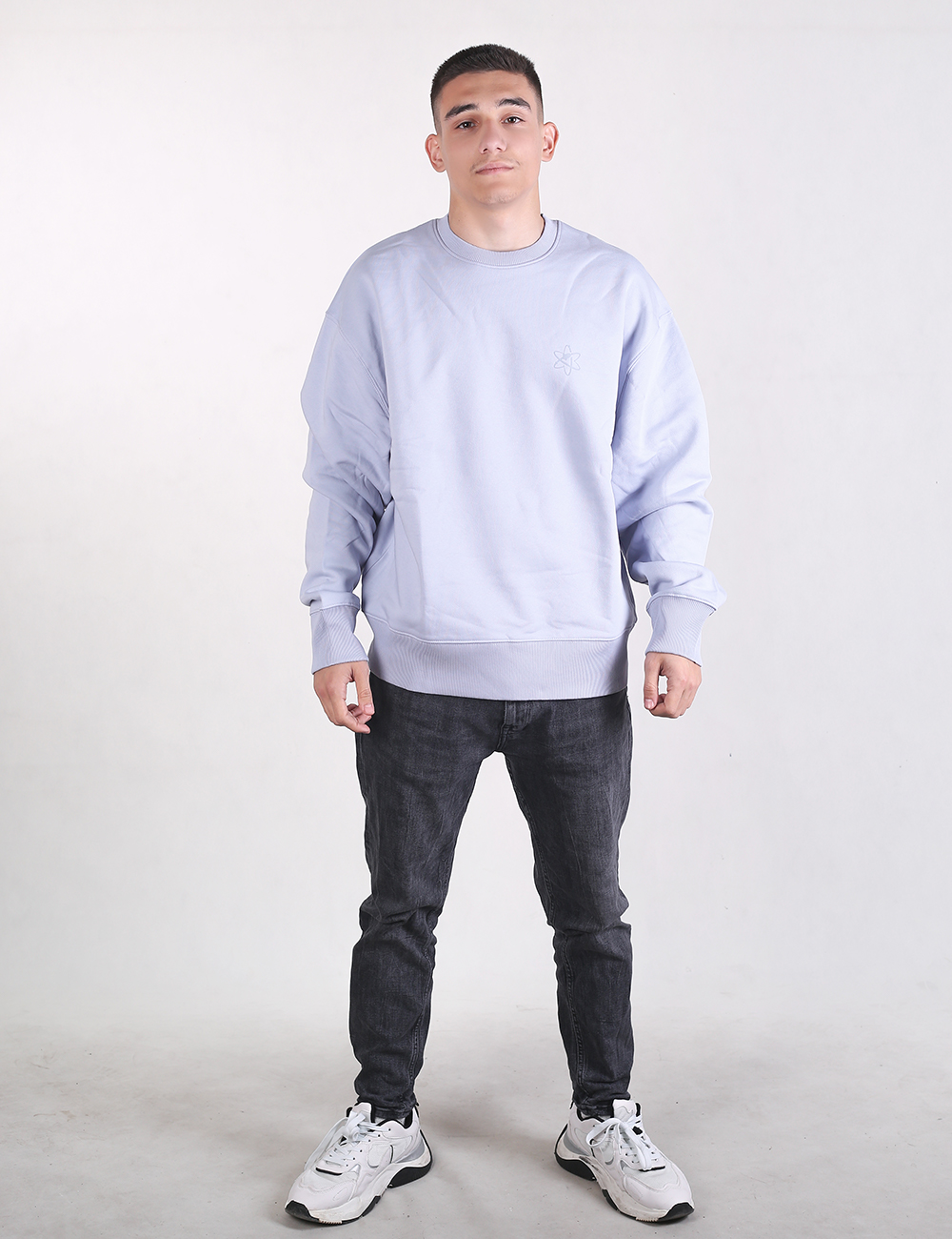 A medium shot of a young man with short dark hair standing against a plain, light grey background. He is facing the camera with a neutral expression and a slight smile. He is wearing an oversized, long-sleeved light blue or lavender crewneck sweatshirt. The sweatshirt has prominent, wide ribbed knit detailing on both the cuffs and the bottom hem. He is also wearing dark grey or black textured jeans, which are visible from the waist down.