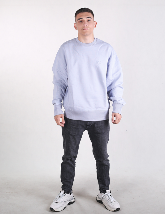A medium shot of a young man with short dark hair standing against a plain, light grey background. He is facing the camera with a neutral expression and a slight smile. He is wearing an oversized, long-sleeved light blue or lavender crewneck sweatshirt. The sweatshirt has prominent, wide ribbed knit detailing on both the cuffs and the bottom hem. He is also wearing dark grey or black textured jeans, which are visible from the waist down.