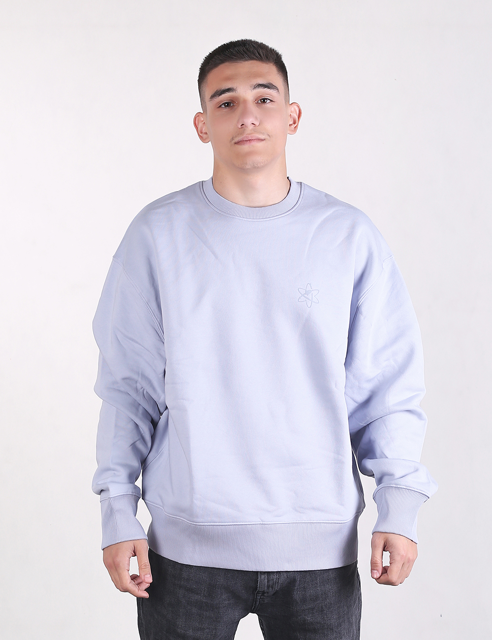 A medium shot of a young man with short dark hair standing against a plain, light grey background. He is wearing an oversized, long-sleeved light blue or lavender crewneck sweatshirt. The sweatshirt has prominent, wide ribbed knit detailing on both the cuffs and the bottom hem. He is also wearing dark grey or black textured jeans, which are visible from the waist down.