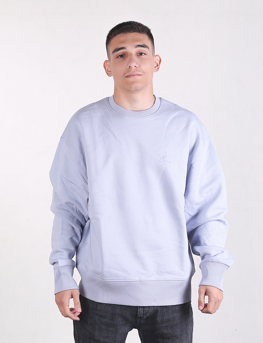 A medium shot of a young man with short dark hair standing against a plain, light grey background. He is wearing an oversized, long-sleeved light blue or lavender crewneck sweatshirt. The sweatshirt has prominent, wide ribbed knit detailing on both the cuffs and the bottom hem. He is also wearing dark grey or black textured jeans, which are visible from the waist down.