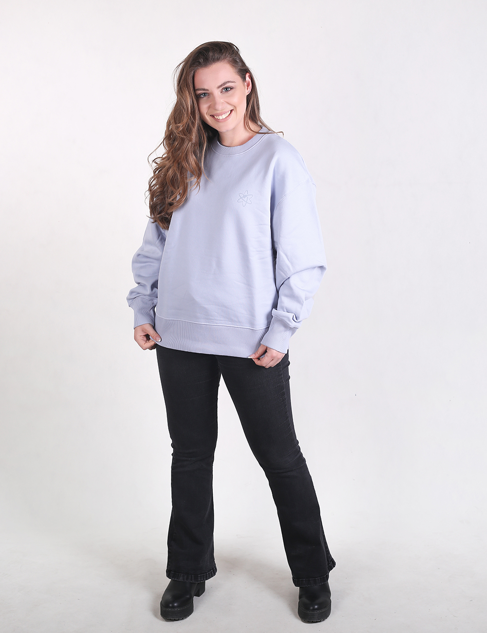 A young woman with long, wavy brown hair stands smiling against a plain white background, wearing an oversized, light blue crewneck sweatshirt with dropped shoulders and ribbed cuffs. She is also wearing black flared denim pants that cover most of her chunky, black platform shoes.