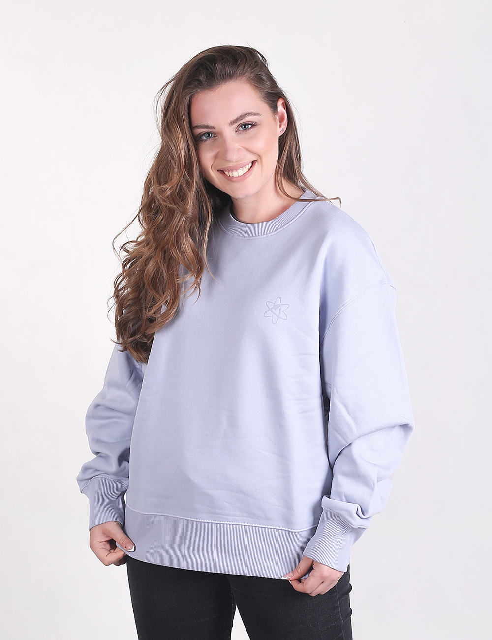 A young woman with long, wavy brown hair stands smiling against a plain white background, wearing an oversized, light blue crewneck sweatshirt with dropped shoulders and ribbed cuffs. She is also wearing black flared denim pants that cover most of her chunky, black platform shoes.