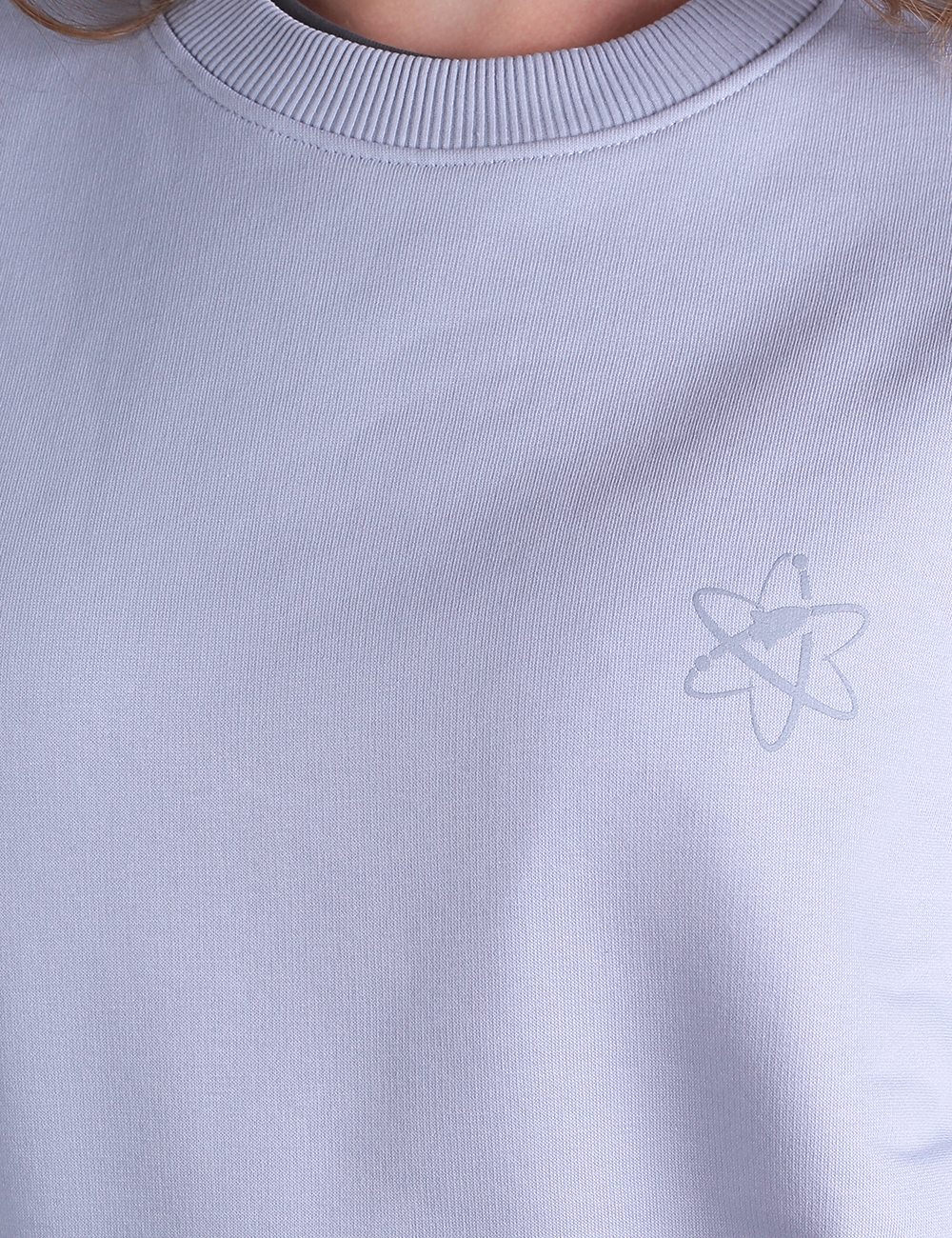 Close-up of a light purple crewneck sweatshirt featuring a ribbed neckline and a subtle, tone-on-tone embroidered atom logo on the upper left chest.