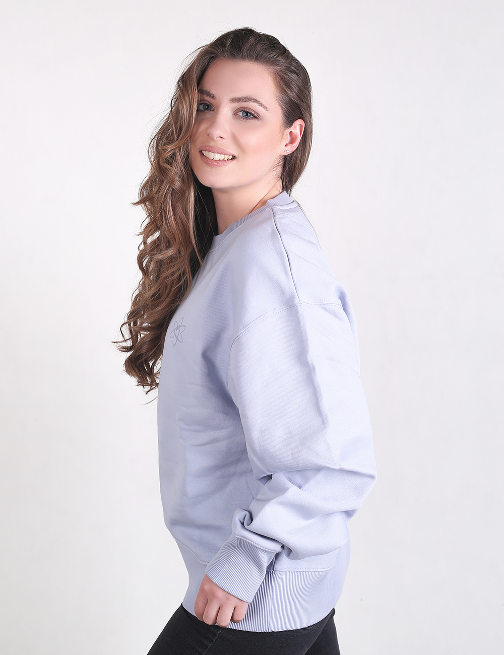 A young woman with long, curly light brown hair smiles while standing in a side-profile pose against a plain, light grey background. She is wearing an oversized, light lilac crewneck sweatshirt with ribbed cuffs and hem, paired with black pants.