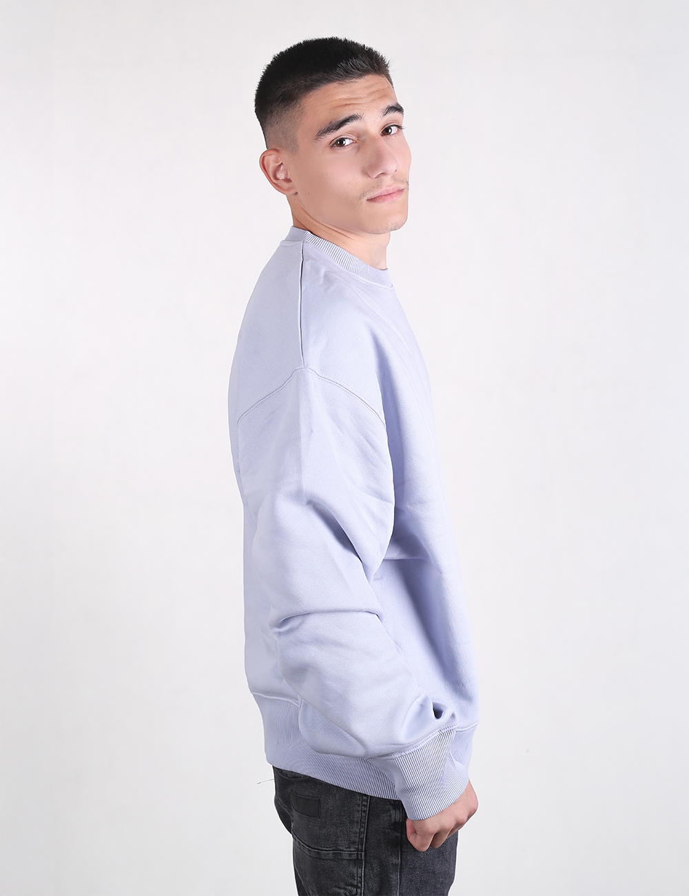 A young man with short cropped dark hair stands in a side profile view against a plain, light grey background, looking back over his shoulder toward the camera. He is wearing an oversized, light lavender-purple crewneck sweatshirt with dropped shoulders and a ribbed hem and cuffs, paired with dark grey or black jeans.